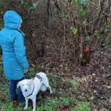 Eine Frau steht mit ihrem Hund vor einem Gebüsch.