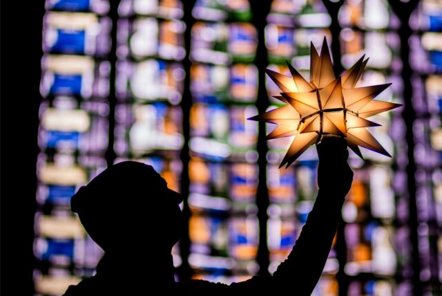 Eine Person hält vor einem Kirchenfenster einen leuchtenden, gebastelten Stern hoch.