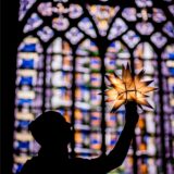 Eine Person hält vor einem Kirchenfenster einen leuchtenden, gebastelten Stern hoch.