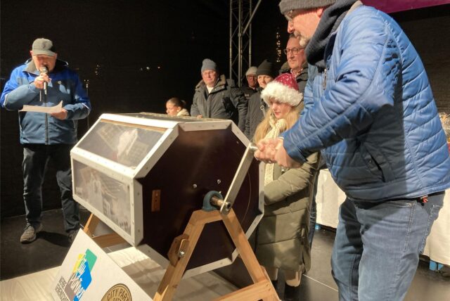 Menschen stehen auf einer weihnachtlich geschmückten Bühne, vor ihnen eine Lostrommel.