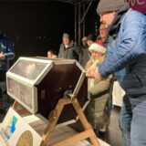 Menschen stehen auf einer weihnachtlich geschmückten Bühne, vor ihnen eine Lostrommel.