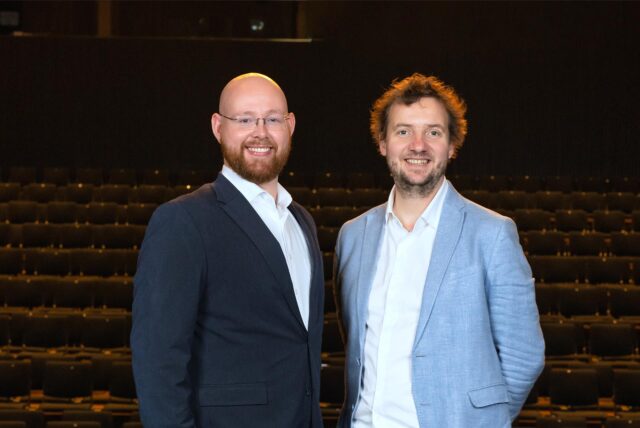Christoph Neumann (links) und Nico Dietrich lächeln in die Kamera. Im Hintergrund sind dunkel Sitze des Theaters in Castrop-Rauxel zu erkennen.