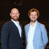 Christoph Neumann (links) und Nico Dietrich lächeln in die Kamera. Im Hintergrund sind dunkel Sitze des Theaters in Castrop-Rauxel zu erkennen.