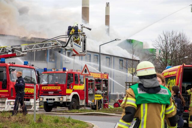 Mehrere Hertener Feuerwehr-Einsatzkräfte bei der Löscharbeit von einer Drehleiter und vom Boden aus. Im Hintergrund quillt dichter Rauch aus einer Gewerbehalle.