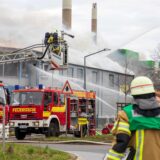 Mehrere Hertener Feuerwehr-Einsatzkräfte bei der Löscharbeit von einer Drehleiter und vom Boden aus. Im Hintergrund quillt dichter Rauch aus einer Gewerbehalle.