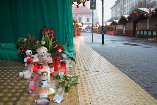 Kerzen, Kuscheltiere und Blumen stehen auf dem Boden an einem Stand auf dem Weihnachtsmarkt. Blumen, Kerzen und Plüschtiere sind als Zeichen des Gedenkens an verscheidenen Orten auf dem Weihnachtsmarkt in Magdeburg und der Innenstadt niedergelegt.