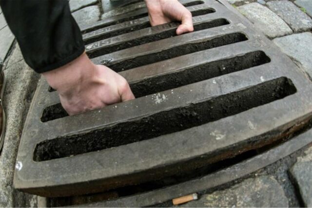 Zwei Männerhände greifen in einen Gullydeckel und sind im Begriff, diesen aus einem Schacht zu heben. (Symbolbild)