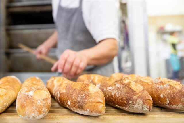 Mehrere Baguette-Brote liegen auf einem Brett. Im Hintergrund sind ein Bäcker und ein Ofen zu sehen.
