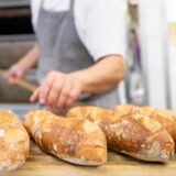 Mehrere Baguette-Brote liegen auf einem Brett. Im Hintergrund sind ein Bäcker und ein Ofen zu sehen.