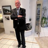 Hubert Schulte-Kemper steht mit verschränkten Armen, klassisch gekleidet im schwarzen Anzug, weißes Hemd, hellblaue Krawatte, in seinem Haus in Alt-Marl.