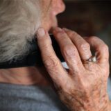 Eine Seniorin mit weißem Haar und grauem Pullover beim Telefonieren, von hinten fotografiert. (Symbolfoto)