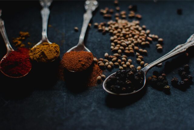 Vier Teelöffel auf einem Tisch, auf denen sich verschiedene Gewürzpulver befinden: Paprika, Curry, Kreuzkümmel und schwarzer Pfeffer.