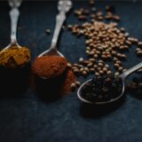 Vier Teelöffel auf einem Tisch, auf denen sich verschiedene Gewürzpulver befinden: Paprika, Curry, Kreuzkümmel und schwarzer Pfeffer.