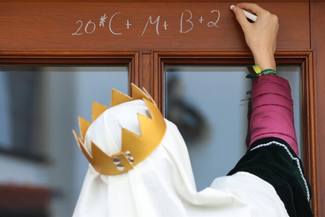 Ein Sternsinger schreibt an Heilige Drei Könige mit Kreide den Segenspruch „C+M+B“ an eine Haustür.