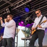 Zwei Männer in weißen Hemden stehen auf einer Bühne. Einer von ihnen singt und der andere spielt Gitarre.