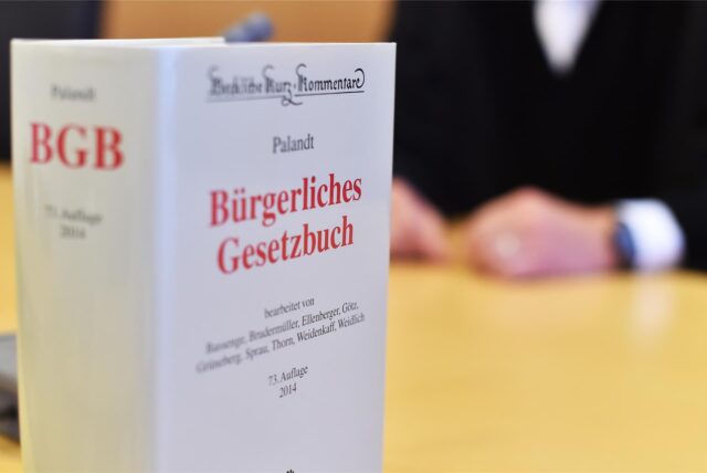 An der Richterbank eines Sitzungssaals beim Oberlandesgericht (OLG) sitzt ein Richter mit Richterrobe, davor steht ein Bürgerliches Gesetzbuch