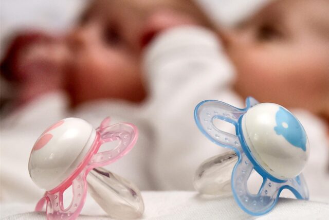 Zwei Schnuller, einer in Rosa und einer in Blau, liegen auf einem Kissen. Im Hintergrund des Bildes sind unscharf zwei Babys zu sehen.
