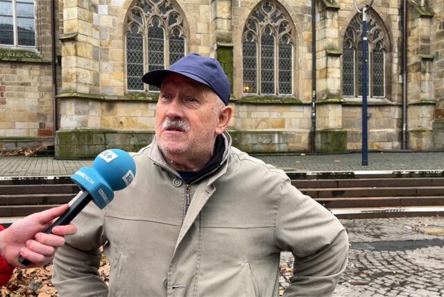 Ein Mann mit einer blauen Kappe und einer hellen Jacke spricht vor einer Kirche in ein blaues RN+-Mikro.