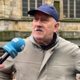 Ein Mann mit einer blauen Kappe und einer hellen Jacke spricht vor einer Kirche in ein blaues RN+-Mikro.