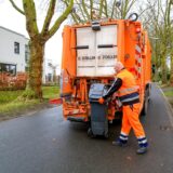 Ein Mitarbeiter der Kommunalen Servicebetriebe Recklinghausen steht hinter einem orangefarbenen Müllauto und entsorgt Müll aus einer schwarzen Mülltonne.