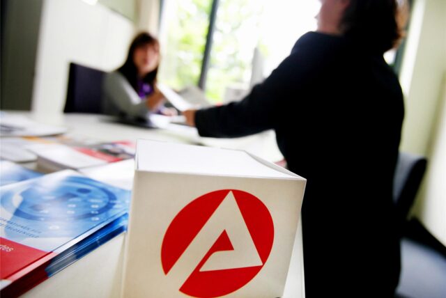 Eine Frau steht an einem Schreibtisch, der das Logo der Agentur für Arbeit trägt, und reicht einer Sachbearbeiterin einen Zettel.