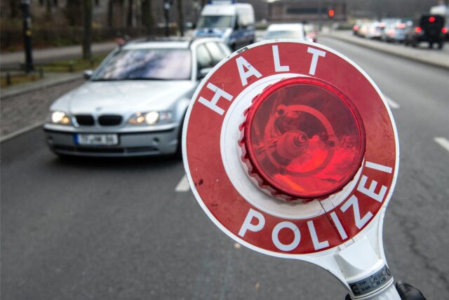 „Halt Polizei“ steht auf der Kelle eines Beamten, im Hintergrund ist ein silberfarbenes Auto zu sehen.