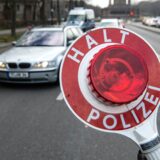 „Halt Polizei“ steht auf der Kelle eines Beamten, im Hintergrund ist ein silberfarbenes Auto zu sehen.