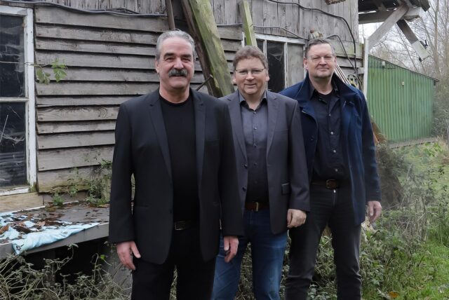 Drei Männer, die Musikgruppe „The Groove Counts“, steht in einem Garten vor einer Holzhütte.