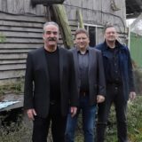 Drei Männer, die Musikgruppe „The Groove Counts“, steht in einem Garten vor einer Holzhütte.