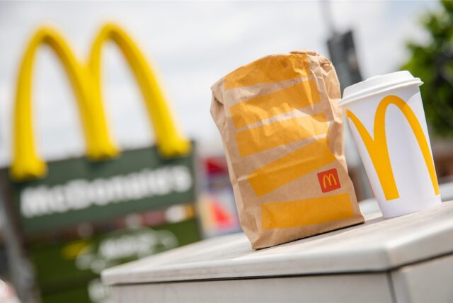 Symbolbild zeigt eine McDonald’s-Tüte und einen Trinkbecher im Vordergrund einer Filiale der Fast-Food-Kette.