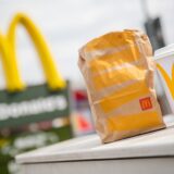 Symbolbild zeigt eine McDonald’s-Tüte und einen Trinkbecher im Vordergrund einer Filiale der Fast-Food-Kette.