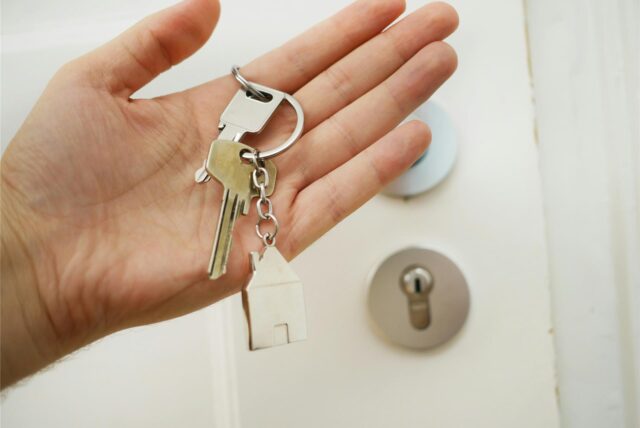 Eine Hand hält einen Schlüssel mit einem Schlüsselanhänger in Form eines Hauses, dahinter ist eine Haustür zu sehen.