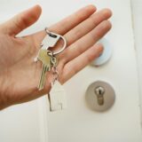 Eine Hand hält einen Schlüssel mit einem Schlüsselanhänger in Form eines Hauses, dahinter ist eine Haustür zu sehen.