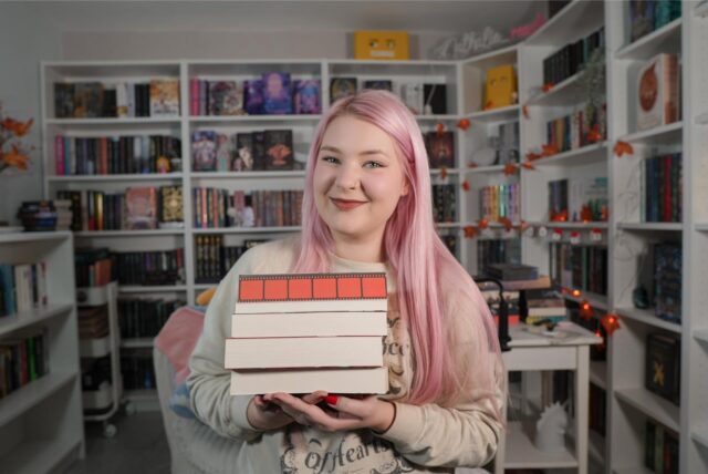 Die Booktok-Influencerin Nathalie Bröske sitzt in ihrem Lesezimmer mit vielen Bücherregalen und hält vier Bücher in die Kamera.