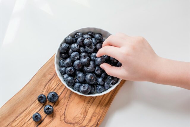 Eine Schale mit Blaubeeren und eine Kinderhand, die nach einer Blaubeere greift.