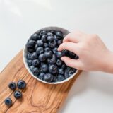 Eine Schale mit Blaubeeren und eine Kinderhand, die nach einer Blaubeere greift.