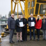 Waltrops Bürgermeister Marcel Mittelbach und LWL-Direktor Dr. Georg Lunemann halten am Schiffshebewerk Henrichenburg in Waltrop die Förderbescheide, die ihnen NRW-Wirtschaftsministerin Mona Neubaur übergeben hatte.