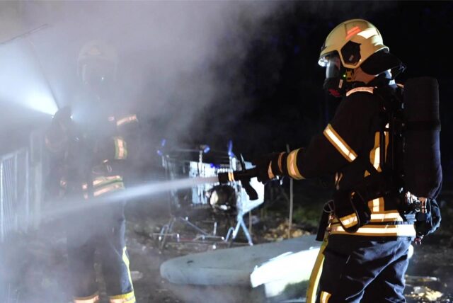 Zwei Feuerwehrleute mit Atemschutzmasken stehen in dichtem Qualm und löschen einen Brand an der Backumer Straße in Herten-Scherlebeck.