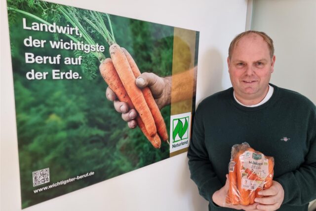 Landwirt Carsten Abenhardt aus Datteln steht vor einem Naturland-Plakat und hält einen Beutel Möhren in den Händen.
