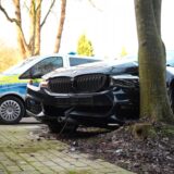 Unfall in Herten: Ein schwarzer BMW ist vor einen Baum geprallt. Dahinter steht ein Polizeiauto.