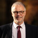 Hertens Bürgermeister Matthias Müller, im dunklen Anzug mit weinroter Krawatte und weißem Hemd, lächelt in die Kamera.