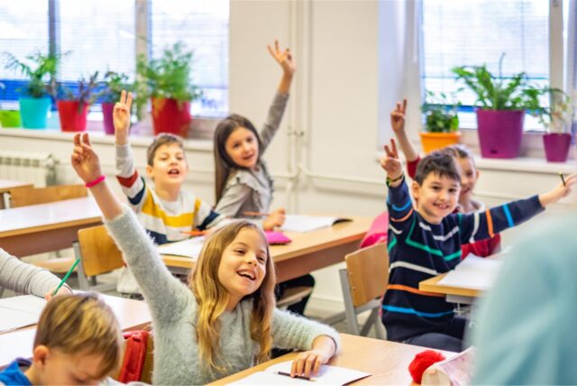 Kinder im Grundschulalter sitzen in einem Klassenzimmer und heben die Arme zum Aufzeigen in die Luft. (Symbolbild)