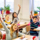 Kinder im Grundschulalter sitzen in einem Klassenzimmer und heben die Arme zum Aufzeigen in die Luft. (Symbolbild)