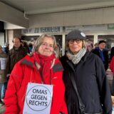 Christiane Rädeld und Ida Dermann stehen unter dem Arkadendach in Marls Hüls. Christiane Rädel trägt ein Schild mit der Aufschrift „Omas gegen Rechts“ um den Hals. Im Hintergrund sind weitere Menschen zu erkennen, manche tragen das gleiche Schild.