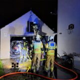 Feuerwehrleute stehen in voller Montur vor einer geöffneten Garage. Darin steht ein mit Löschschaum bedecktes Fahrzeug.