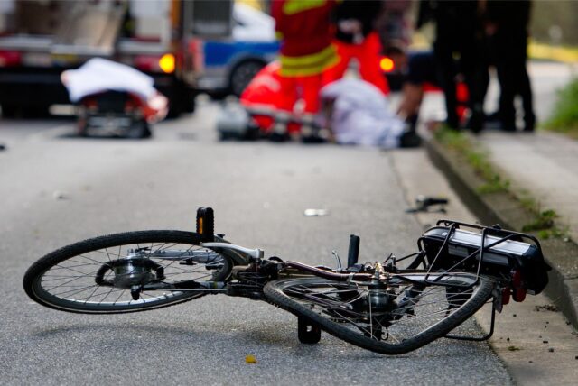 Verunfalltes Fahrrad auf der Straße, im Hintergrund Rettungskräfte.