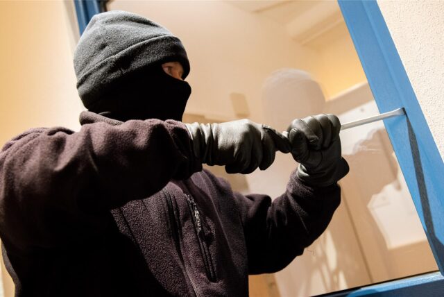 Ein maskierter Mann hebelt ein Fenster mit blauem Rahmen auf.