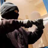 Ein maskierter Mann hebelt ein Fenster mit blauem Rahmen auf.