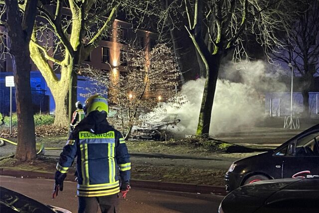 Auf einem Parkplatz brennt ein Auto, davor steht ein Feuerwehrmann.