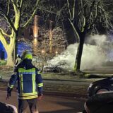 Auf einem Parkplatz brennt ein Auto, davor steht ein Feuerwehrmann.